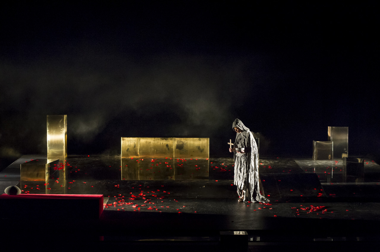 Na kolorowym zdjęciu stojący aktor w pelerynie na scenie pośród scenografii teatralnej spektaklu Irydion.